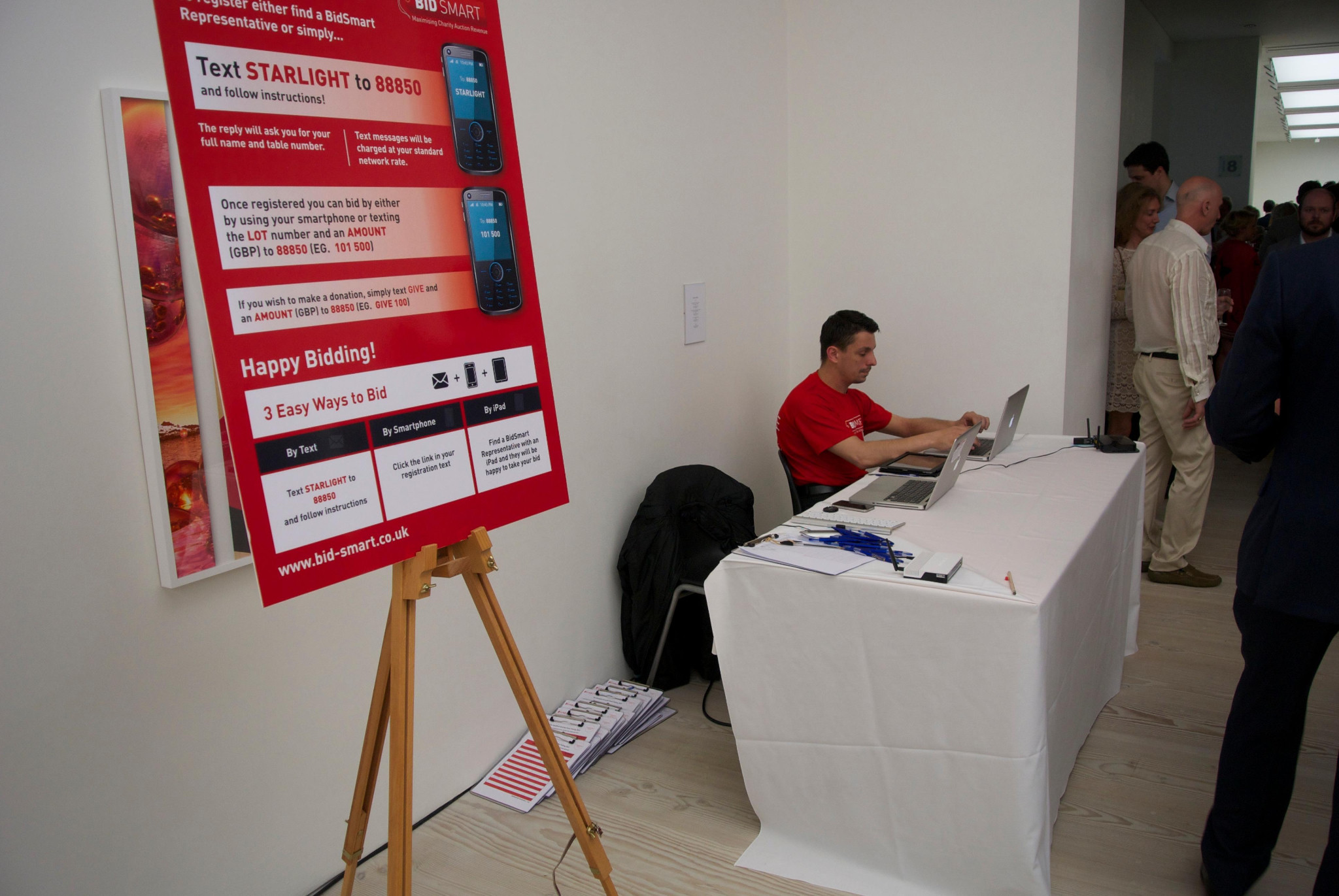 A red BidSmart sign explains text, smartphone and iPad bidding beside a registration table with laptops